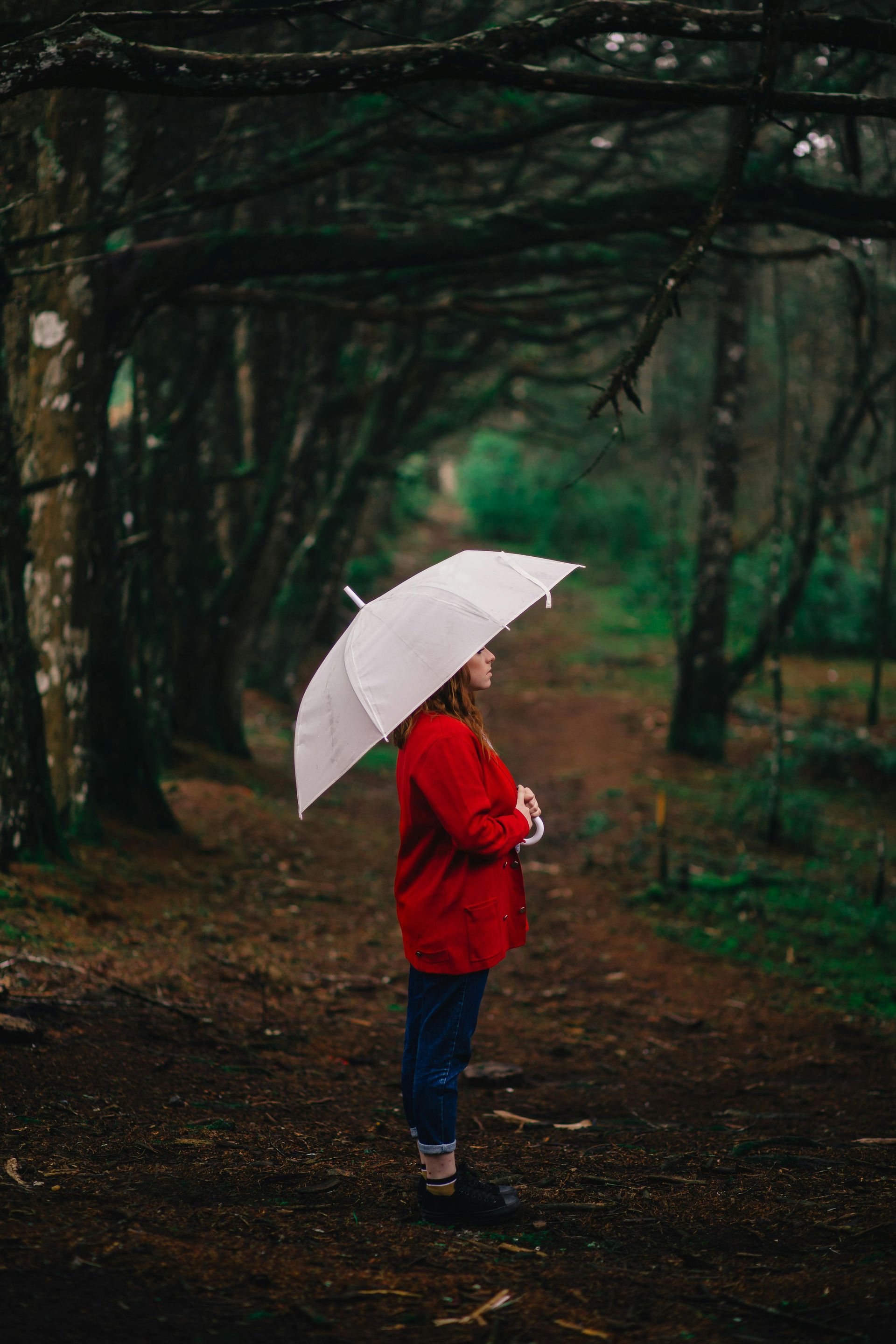 A woman in a red jacket is holding a white umbrella in the woods.