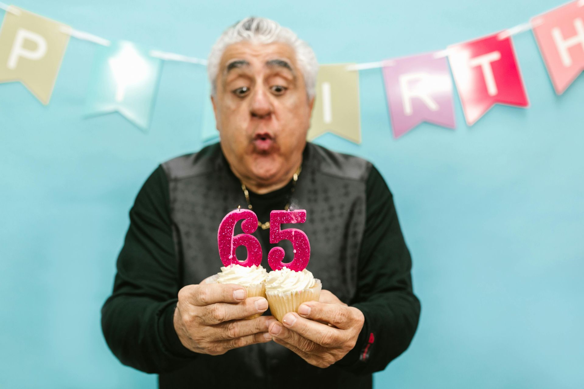 An older man is blowing out candles on two cupcakes.