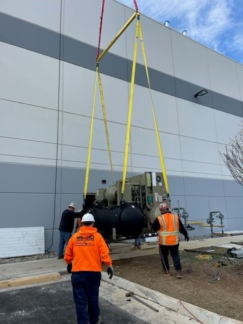 Workers lifting a large generator with yellow straps in front of a gray building.