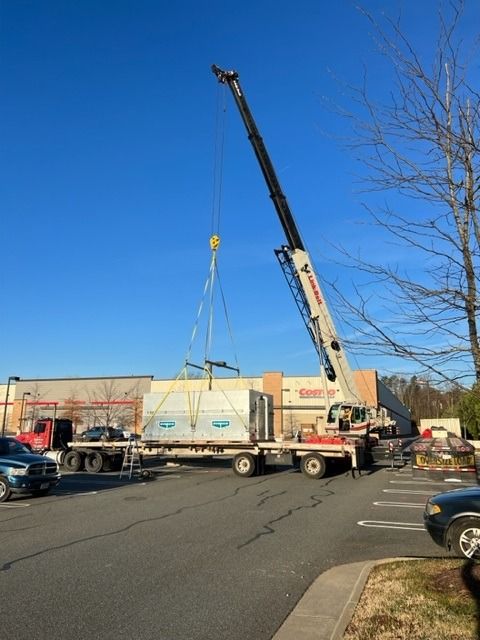 Crane lifting an HVAC unit from a flatbed trailer in a parking lot on a sunny day.