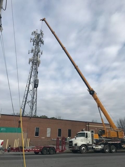 A crane lifting a long beam near a cell tower, in front of a brick building. Sky is cloudy.