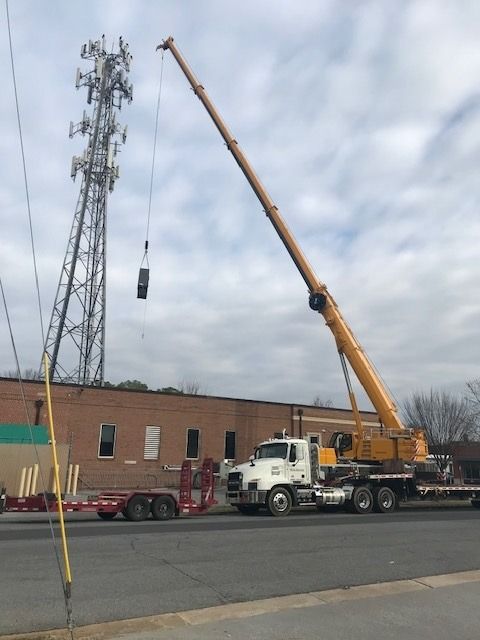 Crane lifting equipment to a cell tower near a brick building.