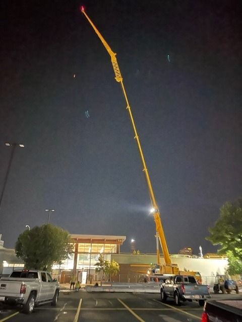 A tall yellow crane with its arm extended upwards at night over a building and parking lot.