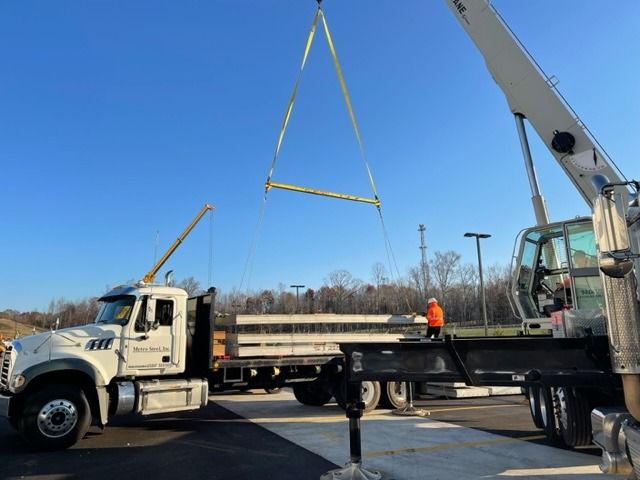 Crane lifting a metal beam from a truck bed. Worker in orange vest observes. Blue sky in the background.