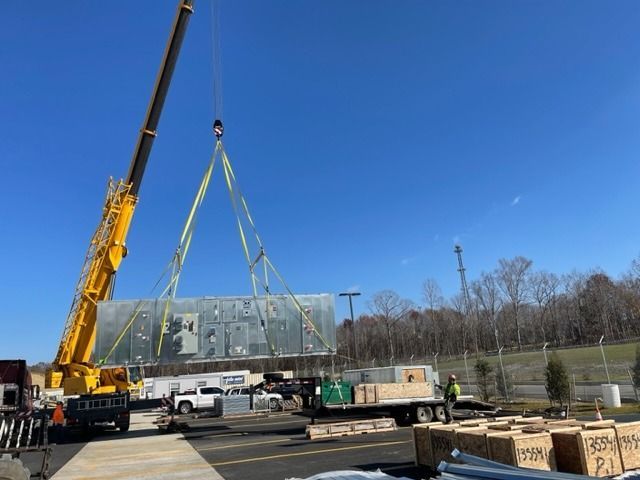 A crane lifts a large, rectangular HVAC unit from a flatbed trailer on a construction site.