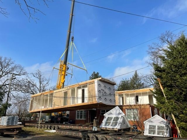 A crane lifts a modular home section onto a foundation. Blue sky.