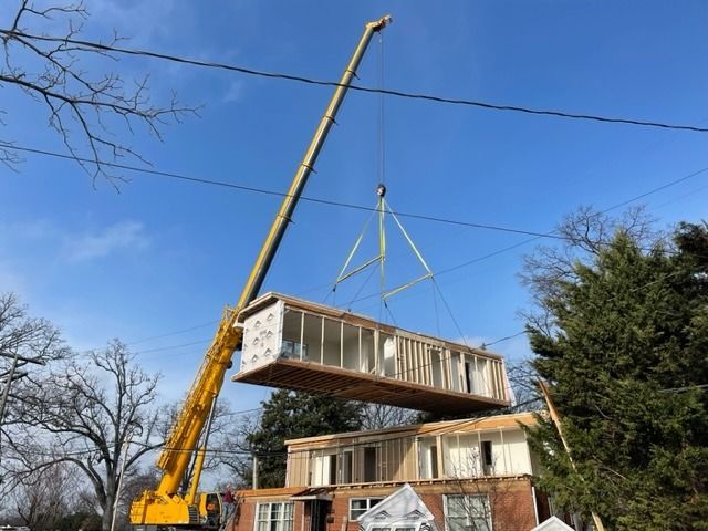 A yellow crane lifting a modular home section over a partially constructed building on a sunny day.