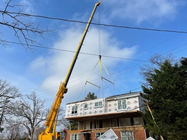 Yellow crane lifting a house's second story. The house has windows and exterior sheathing, under a blue sky.