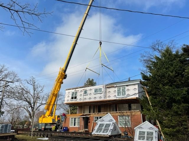 A crane lifting a modular home's second story into place; workers on the roof, sunny day.