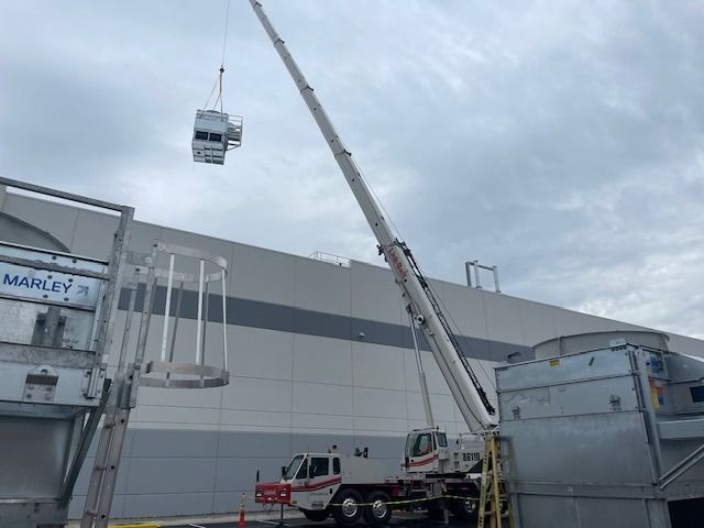 Crane lifting an HVAC unit; other cooling units on the ground; large industrial building in the background.