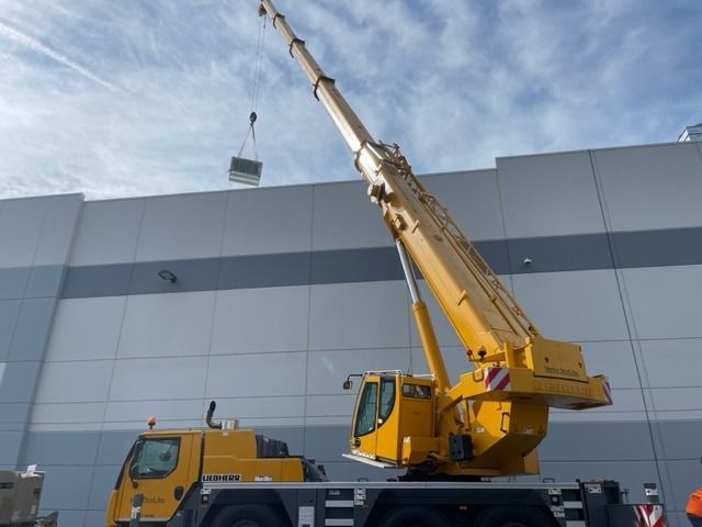 Yellow crane lifting a rectangular object against a white building and blue sky.