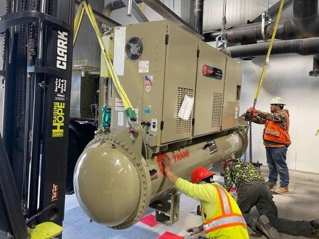 Workers using a forklift to move a large, beige Trane industrial chiller in a building.
