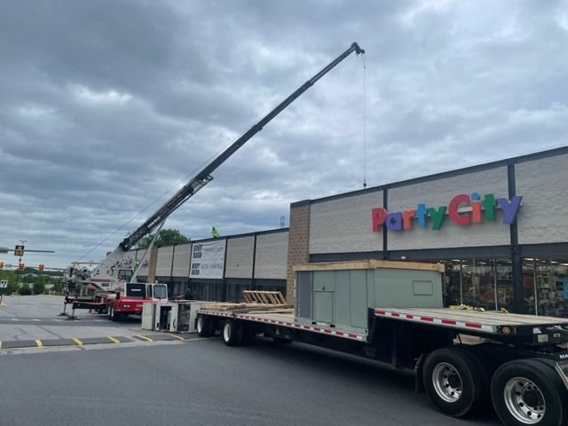 Crane lifting equipment near a Party City store. A flatbed trailer and red truck are present.