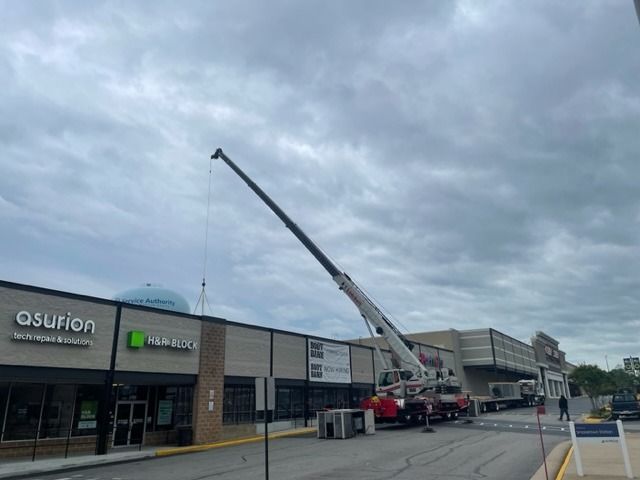 Crane lifting equipment near a row of stores, including an Asurion and a T-Mobile store, under a cloudy sky.