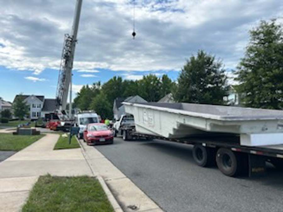 Crane lifting a large rectangular pool shell onto a residential property. A truck, cars, and houses are visible.
