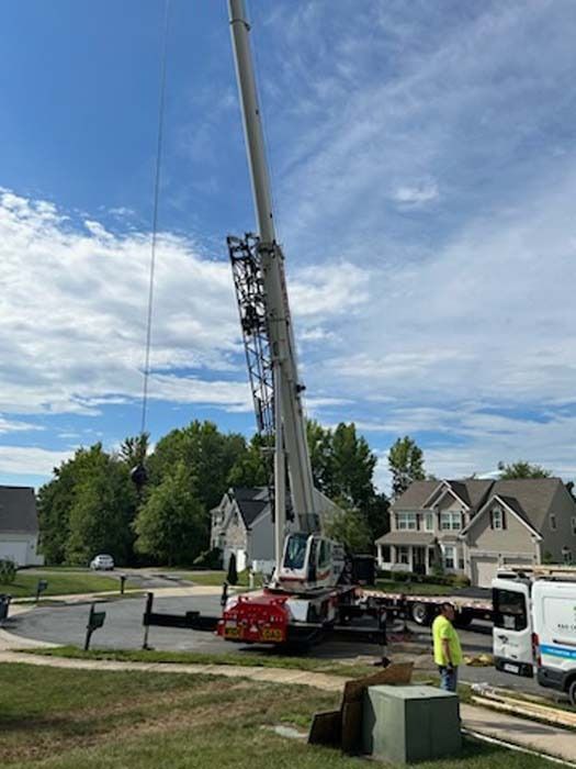A large crane in a suburban setting is lifting something. A worker in a safety vest is standing near it. Blue sky with clouds.