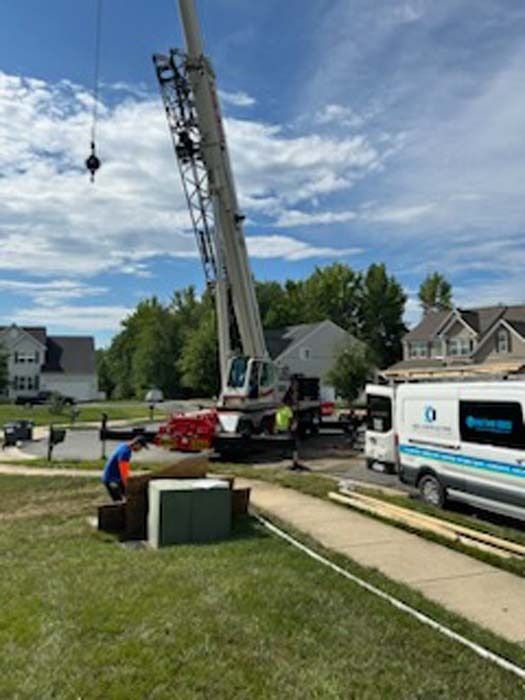Crane lifting a large object near residential houses on a sunny day. Workers and van present.