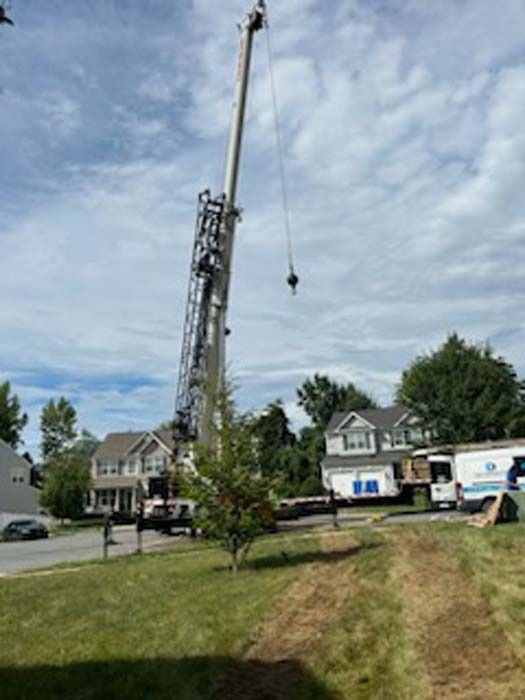 A tall crane over a residential street, lifting something. Houses and trees are in the background. Blue sky.
