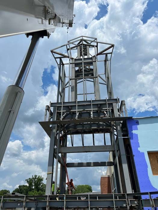 Steel framework of a tower being erected by a crane next to a building. The sky is visible.