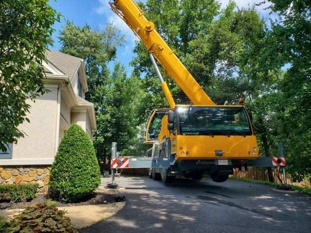 Yellow crane on a driveway, near a house. Crane boom extended upwards. Trees and sky in the background.