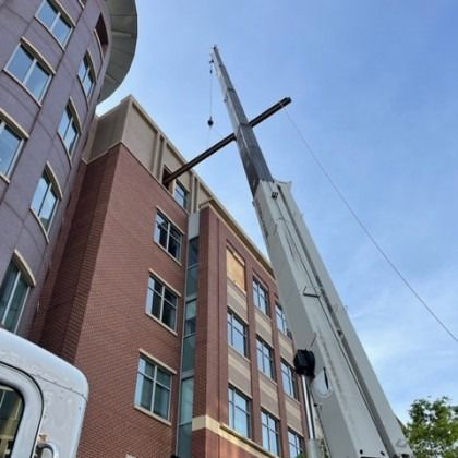 A crane lifting a long metal beam near a multi-story brick building under a blue sky.