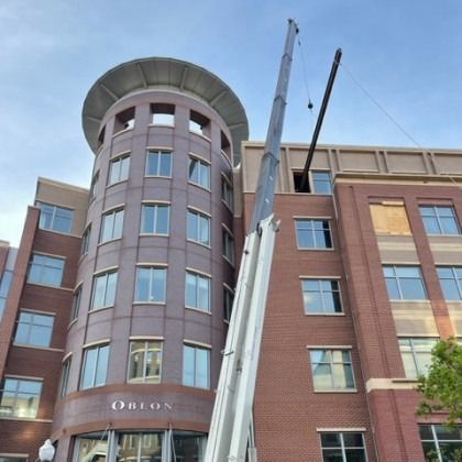 Crane lifting a pipe toward a building, likely for construction. Building is brick with many windows.