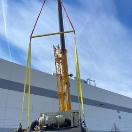 A crane lifting a large, dark mechanical unit with yellow straps against a bright sky.