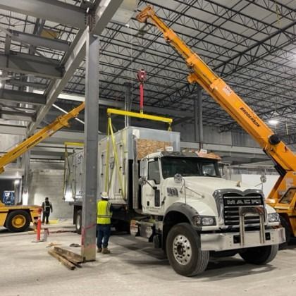 Two yellow cranes lifting a container from a truck inside a large building under construction. Workers in safety vests are present.
