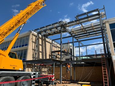 Construction of a steel-framed building addition next to a brick building; crane and workers present.