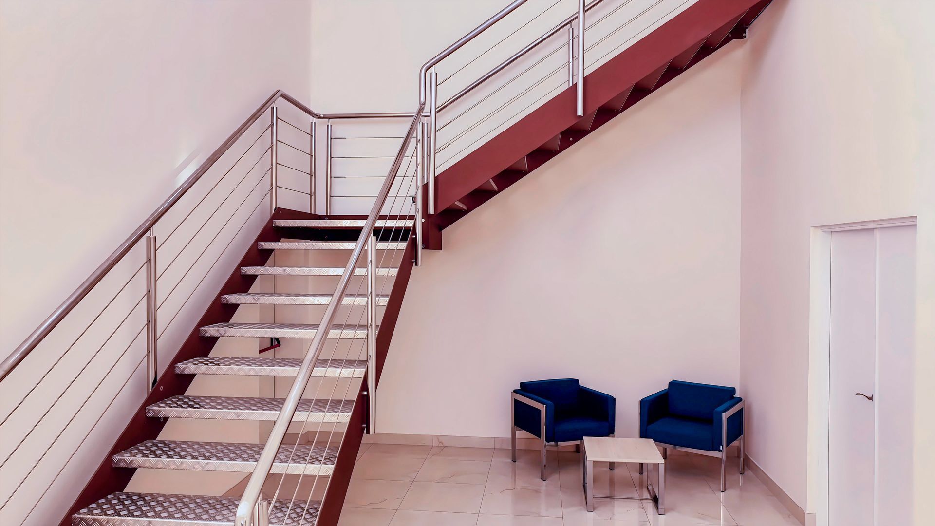 Metal staircase with handrails, leading to an upper level. Two blue armchairs and small table in the waiting area.