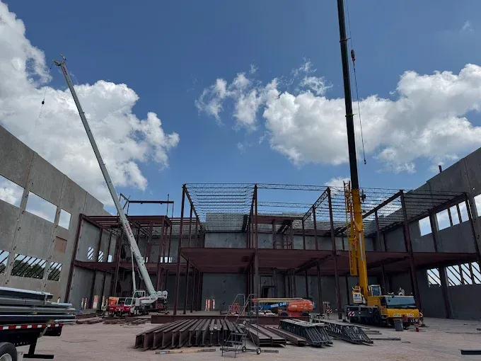 Construction site with steel framework, cranes, and concrete walls against a blue sky.