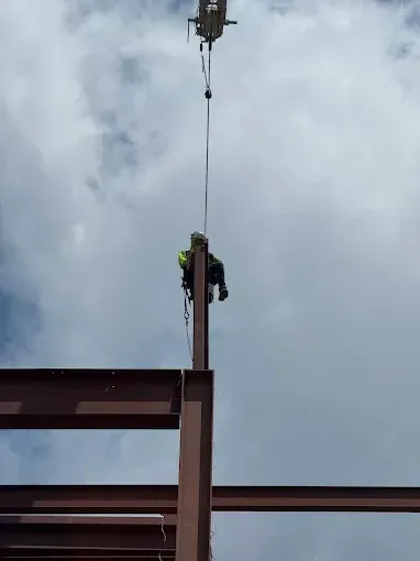 Construction worker on steel beam, suspended by a crane cable against a cloudy sky.