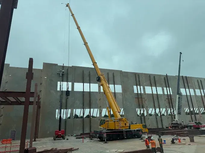 Construction site with a large yellow crane lifting a concrete wall panel. Workers and other machinery are present.