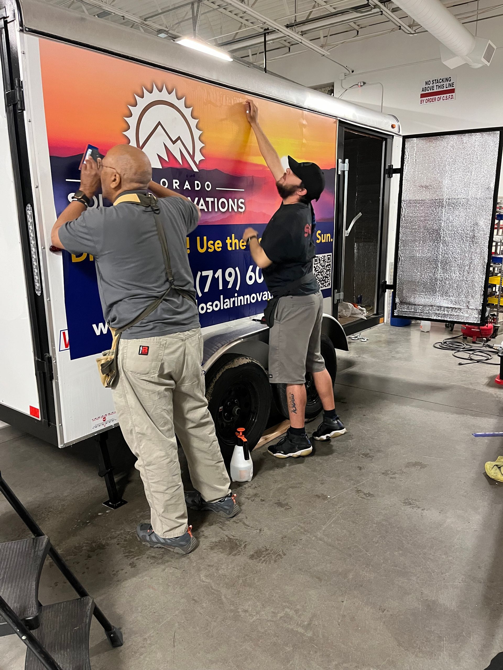 Two men are working on a trailer in a garage installing a vehicle wrap