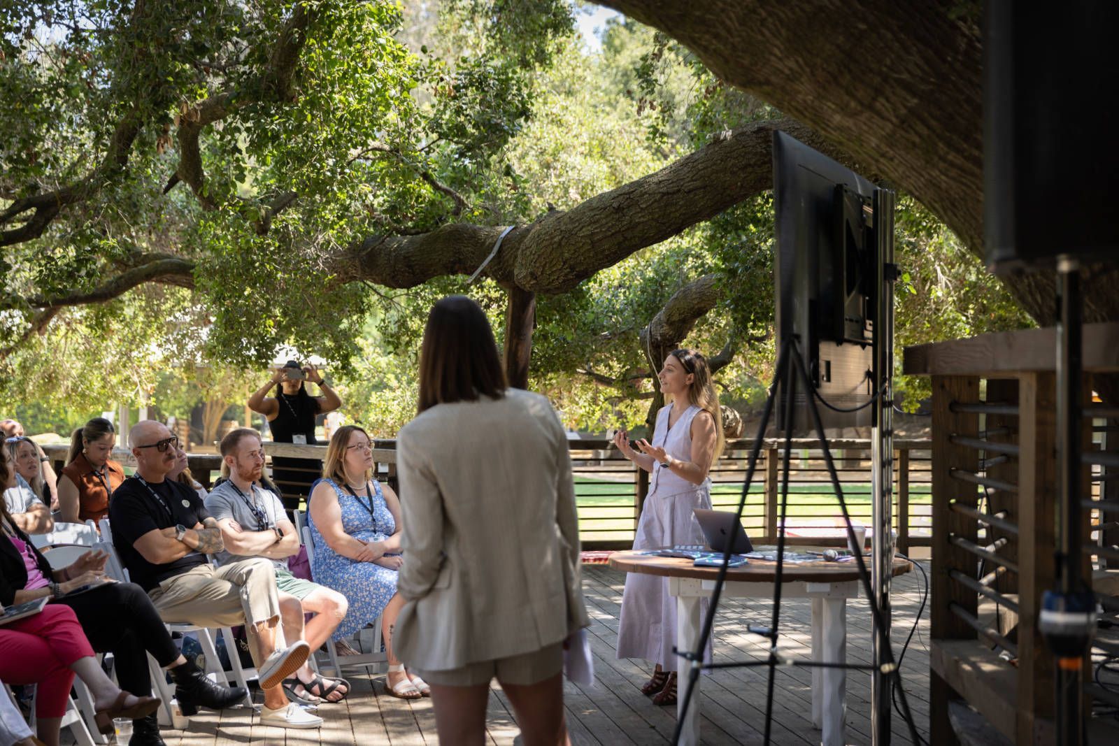 A woman is giving a presentation to a group of people sitting under a tree.