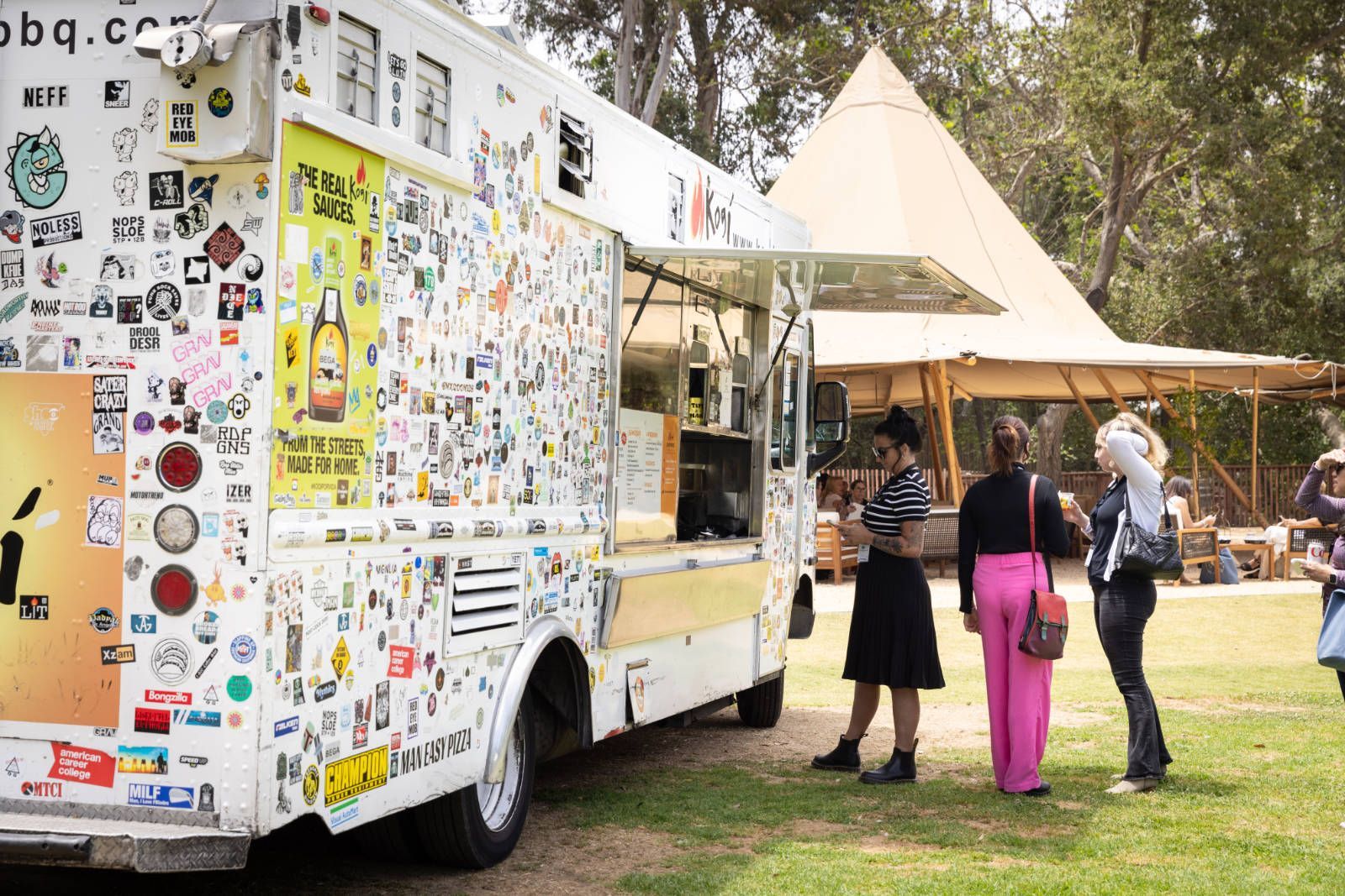 A food truck with a lot of stickers on it is parked in a grassy field.