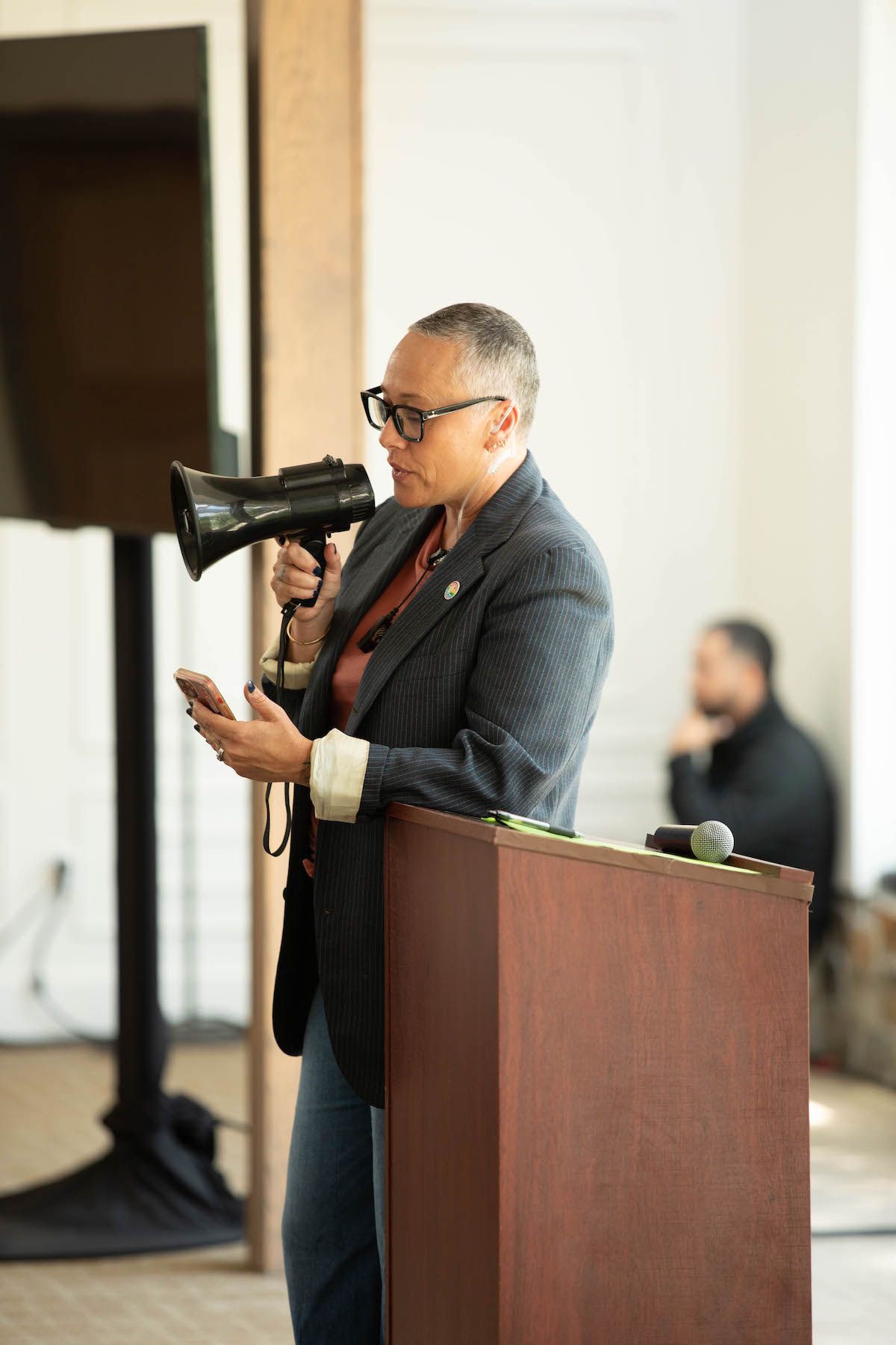 A woman is standing at a podium holding a megaphone and a cell phone.