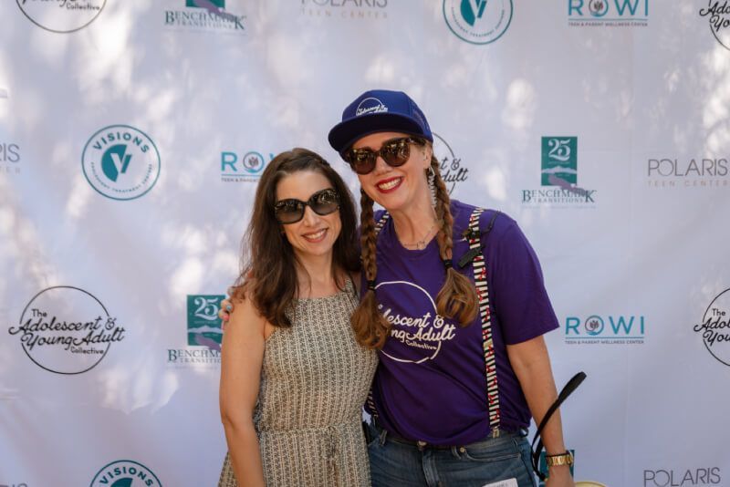 Two women are posing for a picture in front of a wall with logos on it.
