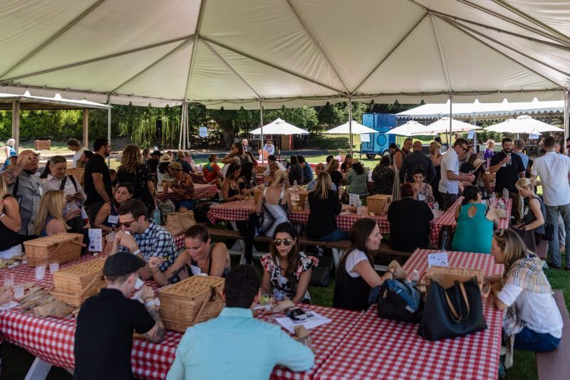 A large group of people are sitting at picnic tables under a tent.