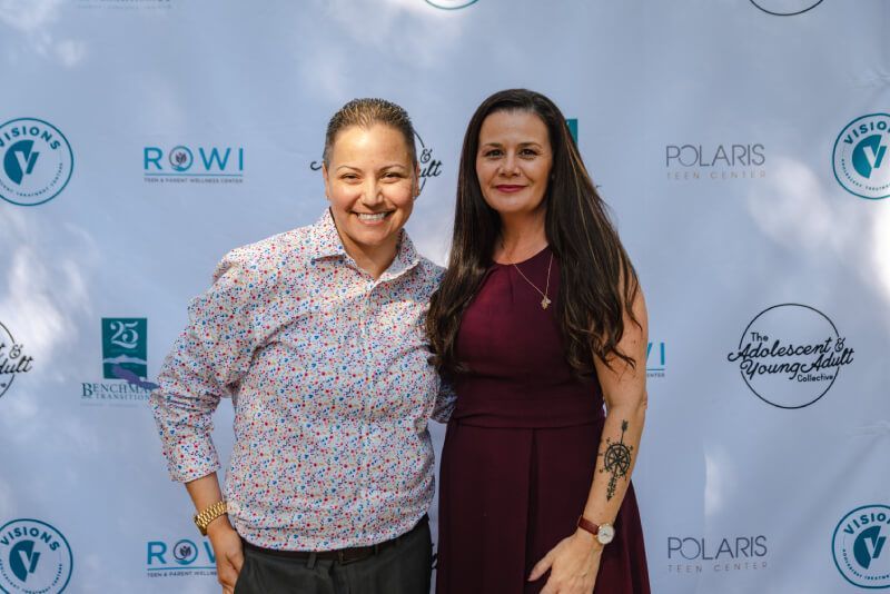 Two women are posing for a picture in front of a white backdrop.
