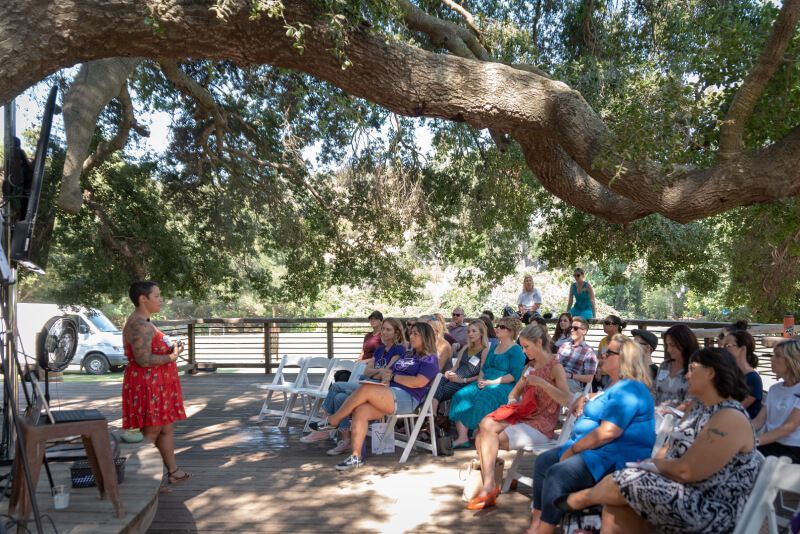 A woman is standing in front of a group of people sitting under a tree.