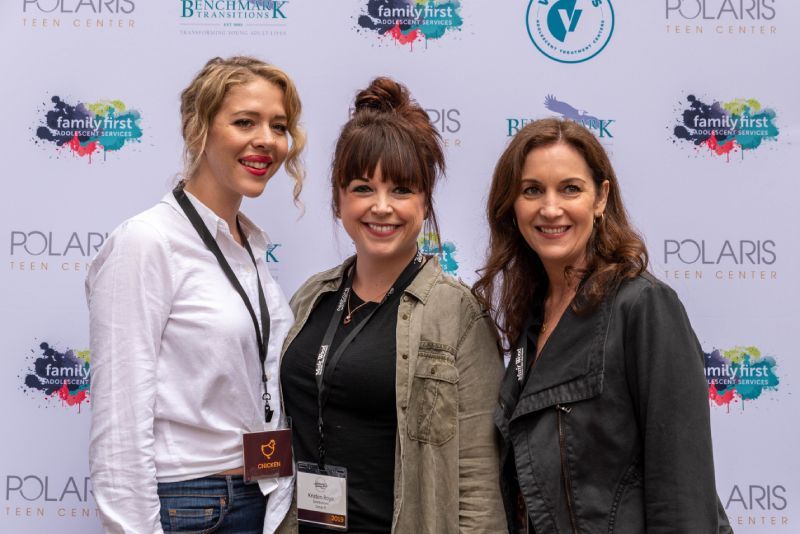 Three women are posing for a picture in front of a wall with logos on it.
