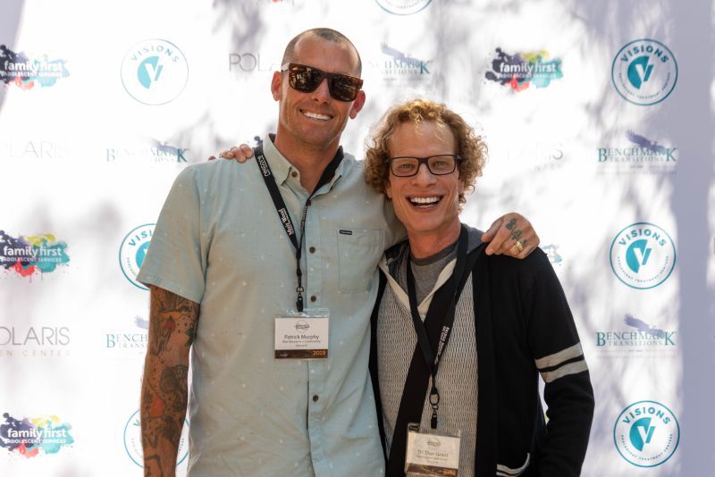 Two men are posing for a picture in front of a purple backdrop.