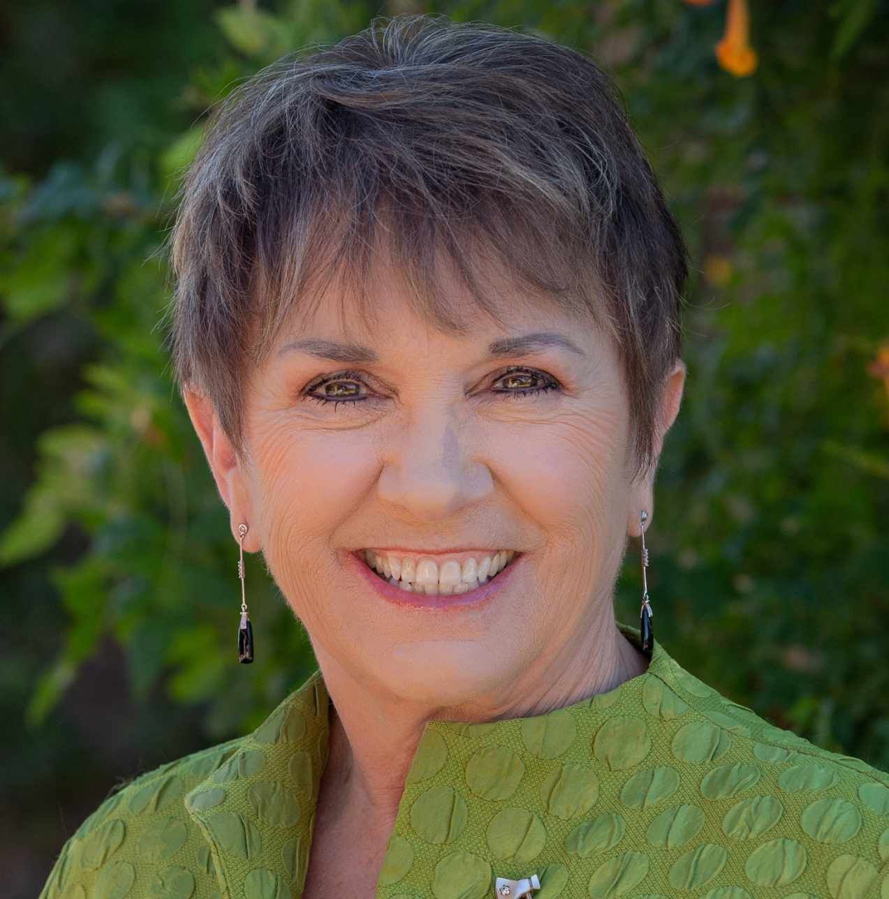 Woman with short brown hair, smiling in a green jacket, outdoor background.