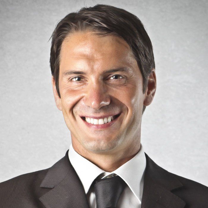 Smiling man in a dark suit, white shirt, and black tie against a light gray background