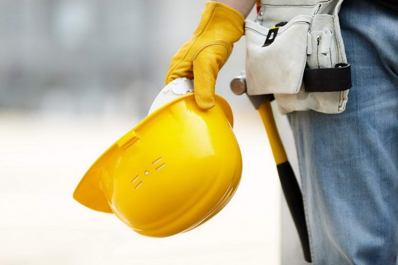 Yellow hard hat held by a gloved construction worker beside a tool belt and jeans