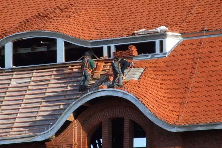 Workers repairing a red-tiled roof beside a dormer window