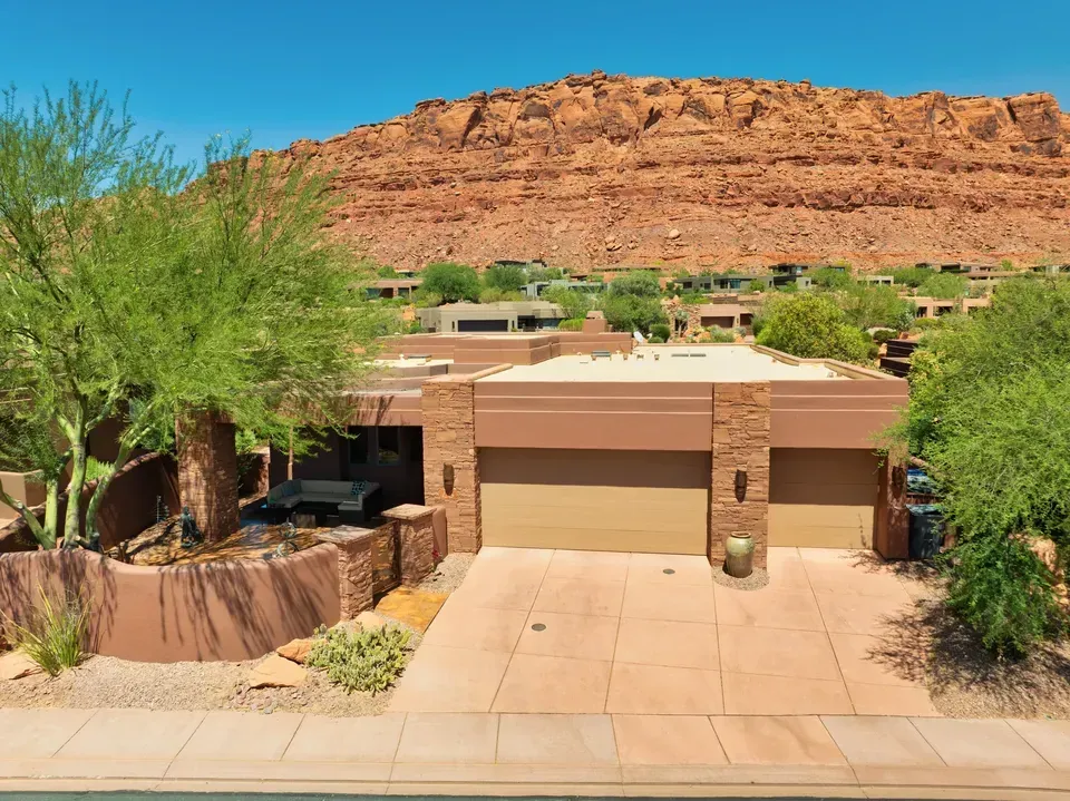 Desert home with beige garage, tan driveway, and red rock cliffs in the background