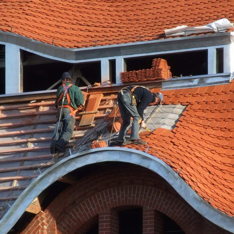 Workers repairing a red-tiled roof on a building, with one standing on a ladder and another kneeling.