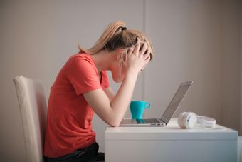Uma mulher está sentada em uma mesa com a cabeça entre as mãos em frente a um laptop.